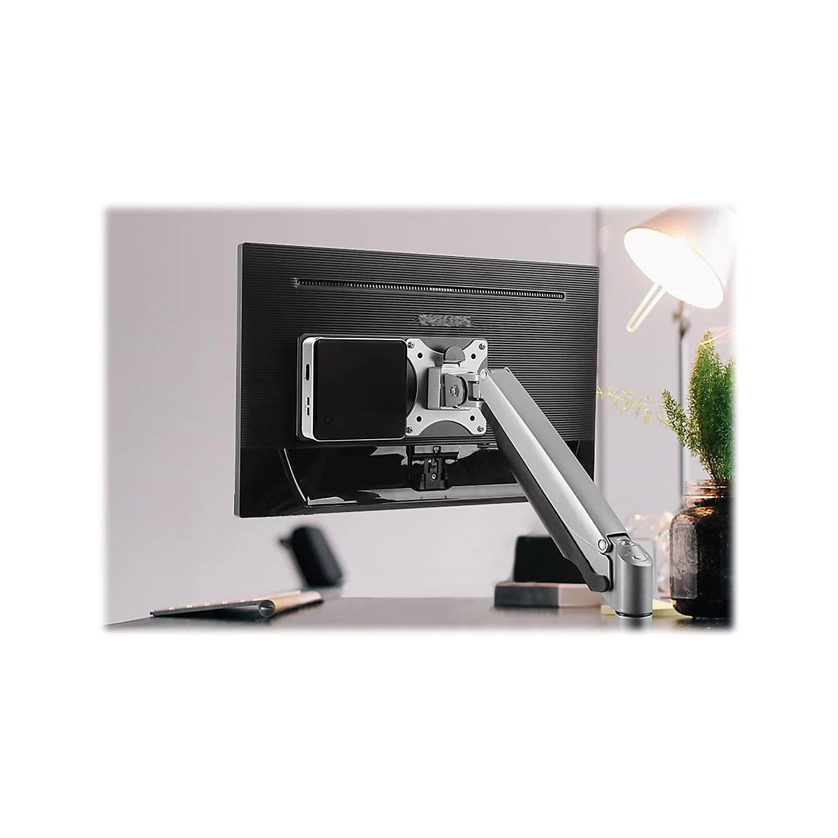 Monitor mit Halterung, montiert an einem Schreibtisch. Im Hintergrund eine Lampe und Pflanzen.