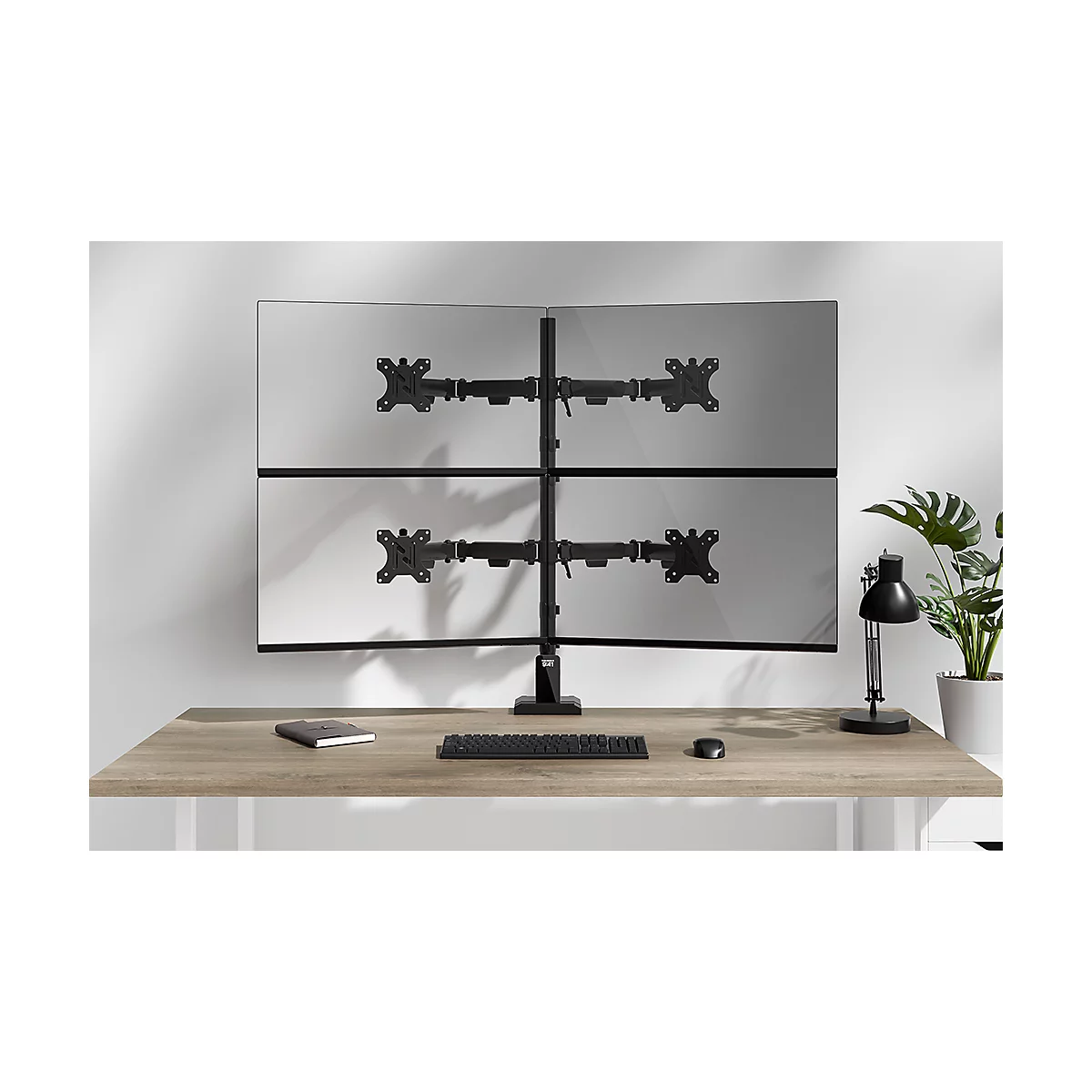 Een monitorstandaard met vier monitoren op een bureau. Toetsenbord, muis, lamp en plant in de foto.
