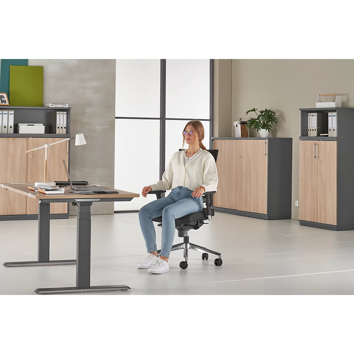Young woman sits in an office chair at a desk. She wears jeans, a cream top, and glasses. Office furniture in the background.