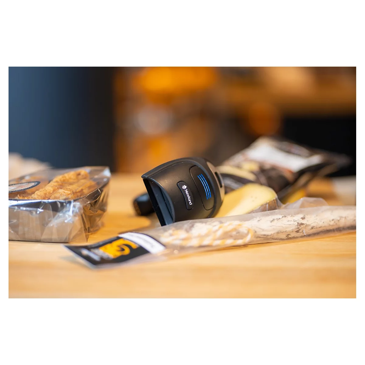 Ein schwarzer Barcodescanner liegt auf einem Tisch neben verpackten Lebensmitteln. Der Scanner ist auf einen Barcode gerichtet.