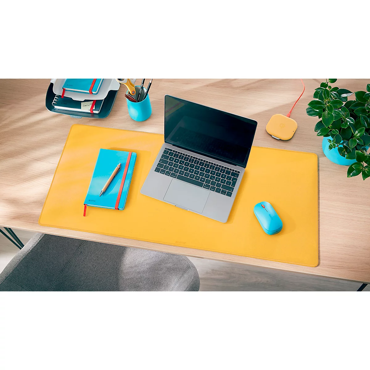 Gelbes Mousepad mit Laptop, Maus und Notizbuch auf hellem Holztisch. Rechts eine Topfpflanze.