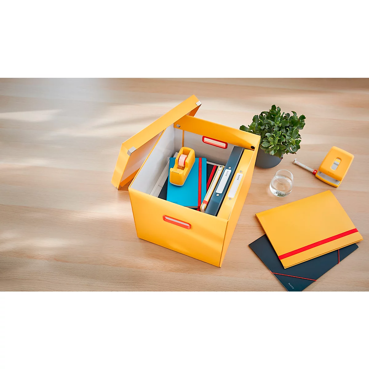 Gelbe Box mit Bürobedarf geöffnet, daneben Notizbücher, Locher und Pflanze.