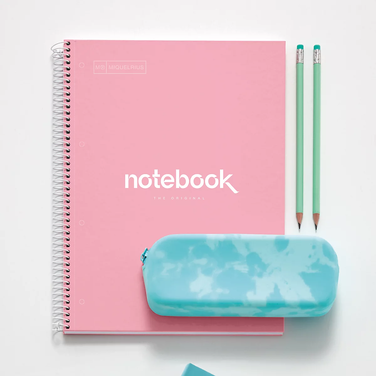 Rosa Notizbuch mit weißem 'Notebook'-Schriftzug, hellgrüne Stifte und türkisgrünes Etui auf weißem Hintergrund.
