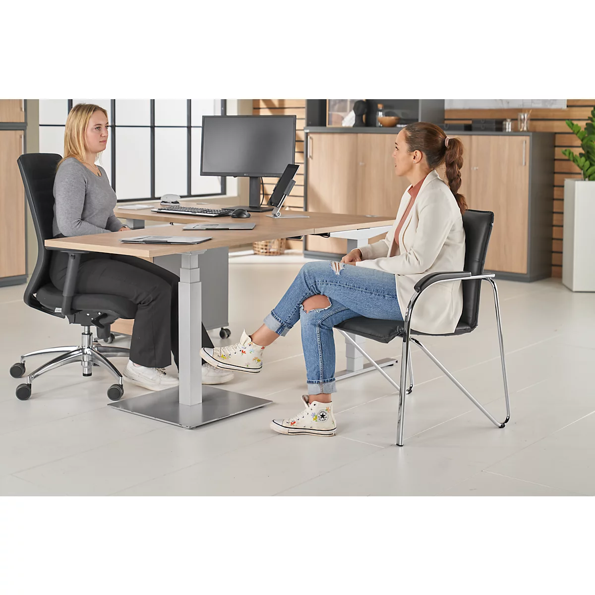 Zwei Frauen sitzen an einem Schreibtisch. Eine trägt einen grauen Pullover, die andere Jeans und Jacke.