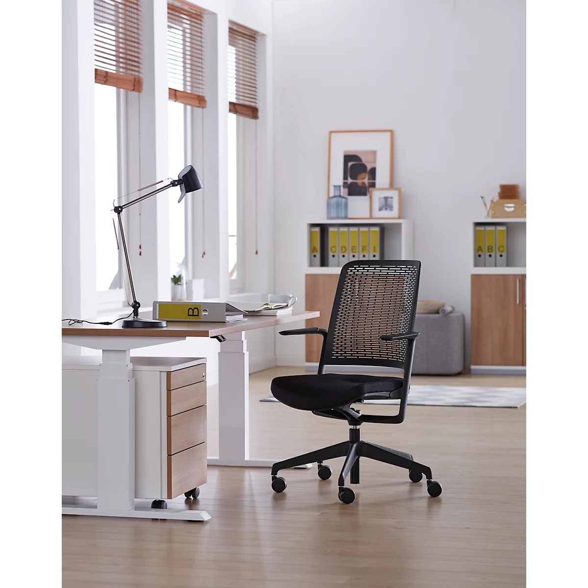 Bureaustoel voor een raam. De stoel heeft een zwart frame en een geweven rugleuning. Het bureau is wit met een houten afwerking.