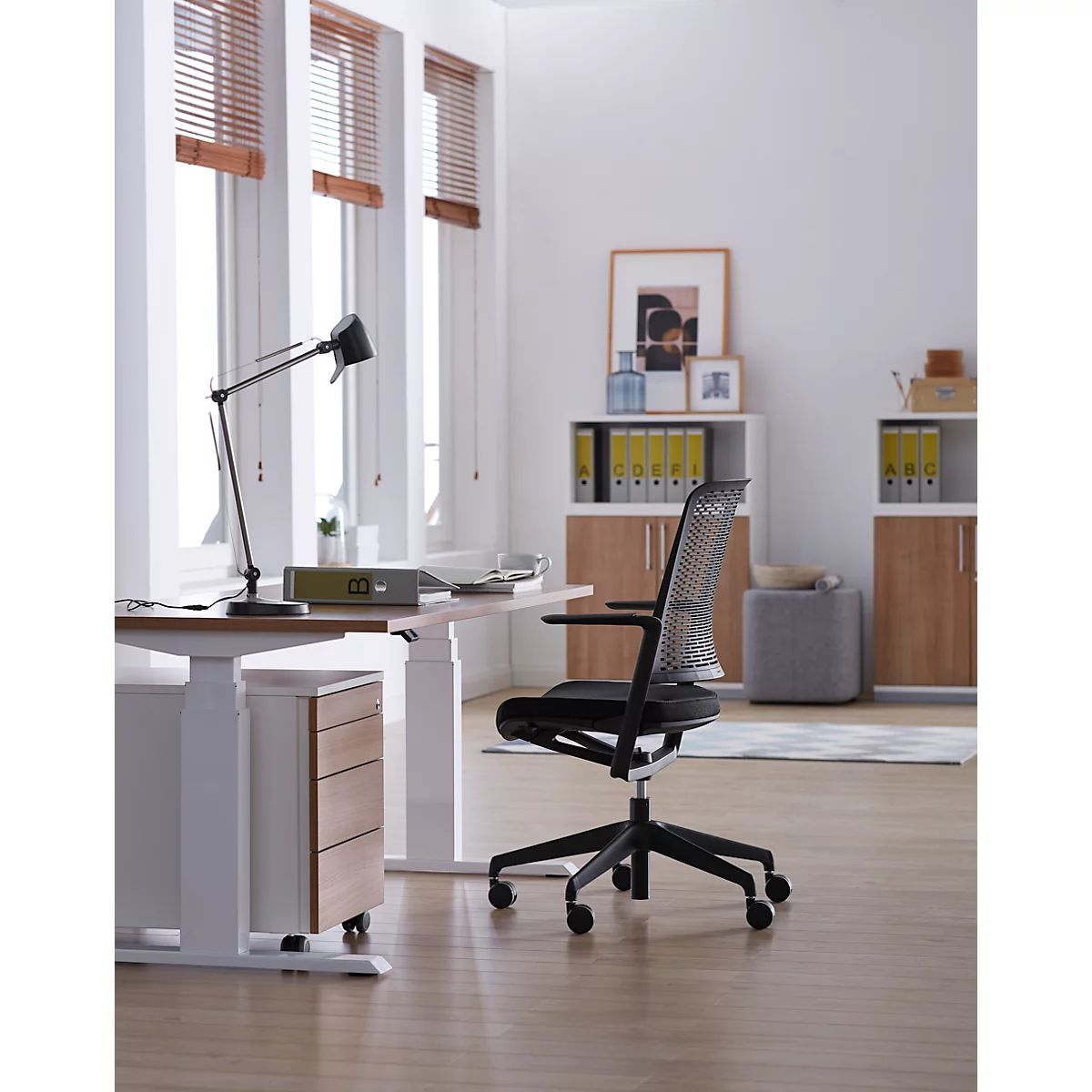 Lichte kantoorruimte met bureau, bureaustoel, kast en ramen met jaloezieën. Een bureau met lamp en opbergruimte. De stoel staat in het midden.