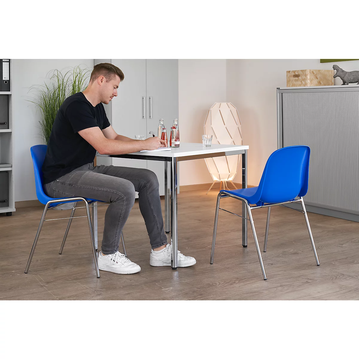 Ein Mann sitzt an einem Tisch, schreibt. Blauer Stuhl, Wasserflasche, Lampe im Hintergrund.