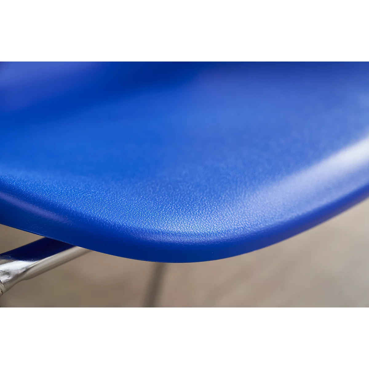 Nahaufnahme eines blauen Stuhlsitzes mit abgerundeter Kante. Chromfarbene Stuhlbeine im unteren Bildbereich.