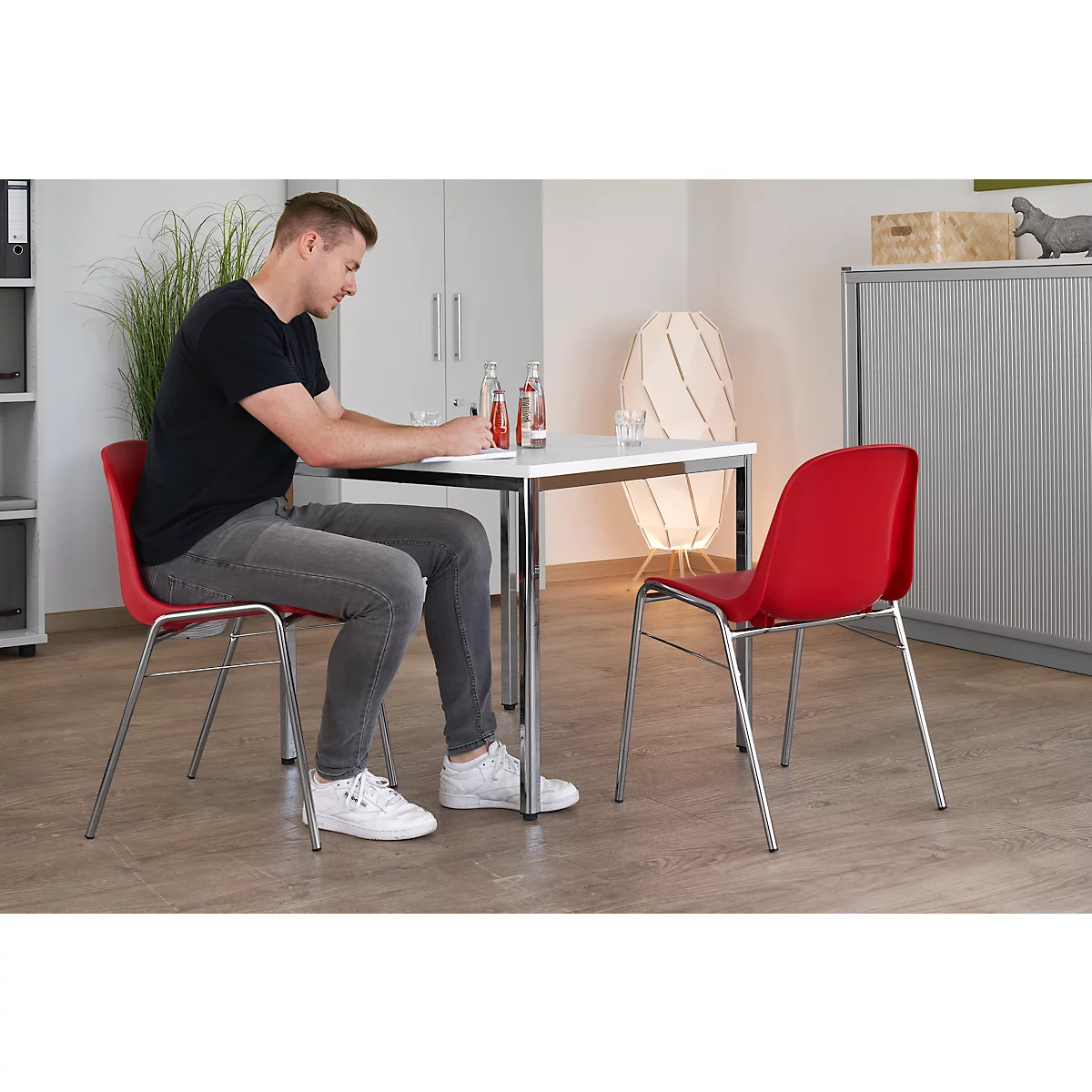 Mann am Tisch, schreibt. Roter Stuhl, Tisch mit Getränken. Helles Büro.