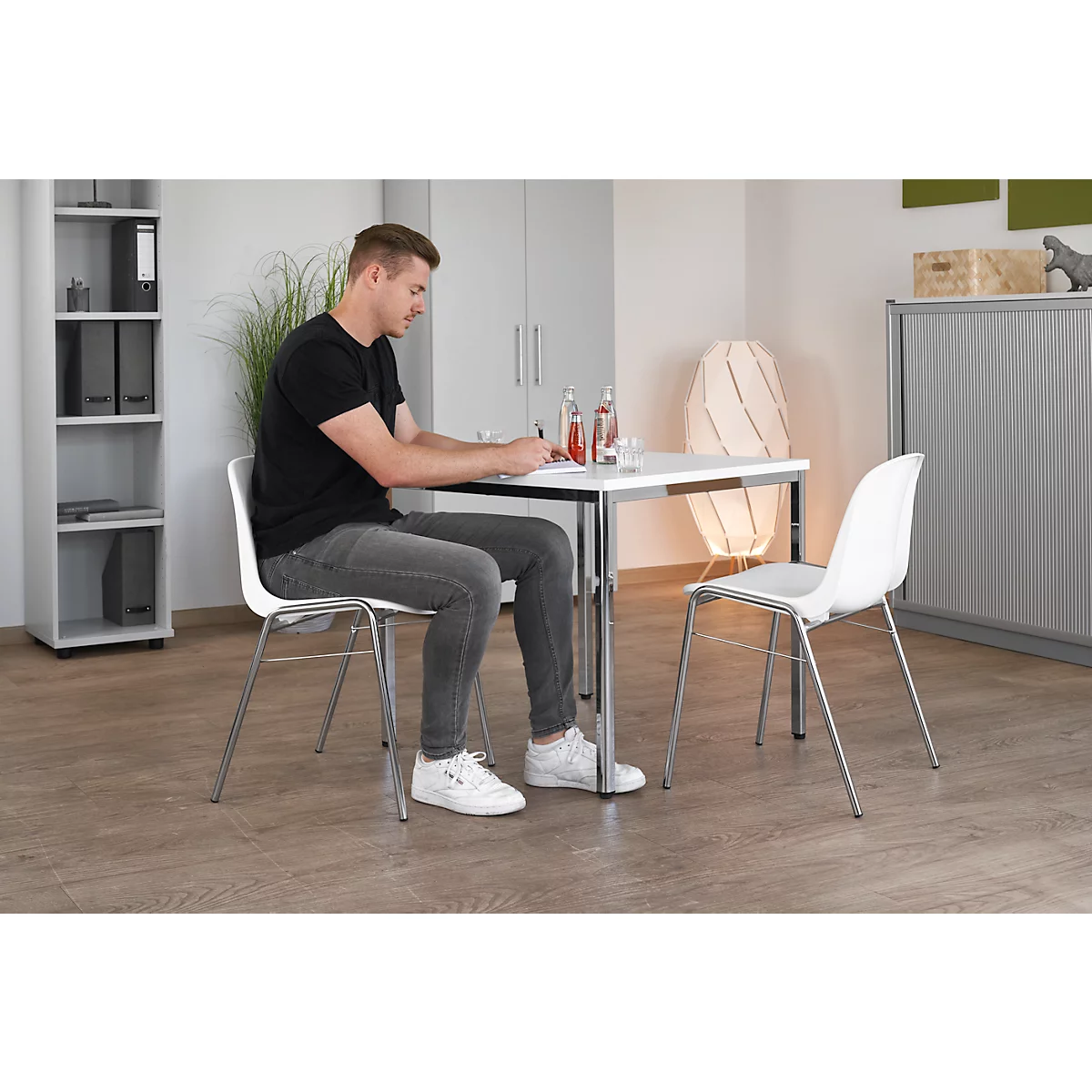 Mann sitzt an einem weißen Tisch, schreibt. Zwei Stühle. Wasserflaschen. Helle Einrichtung.