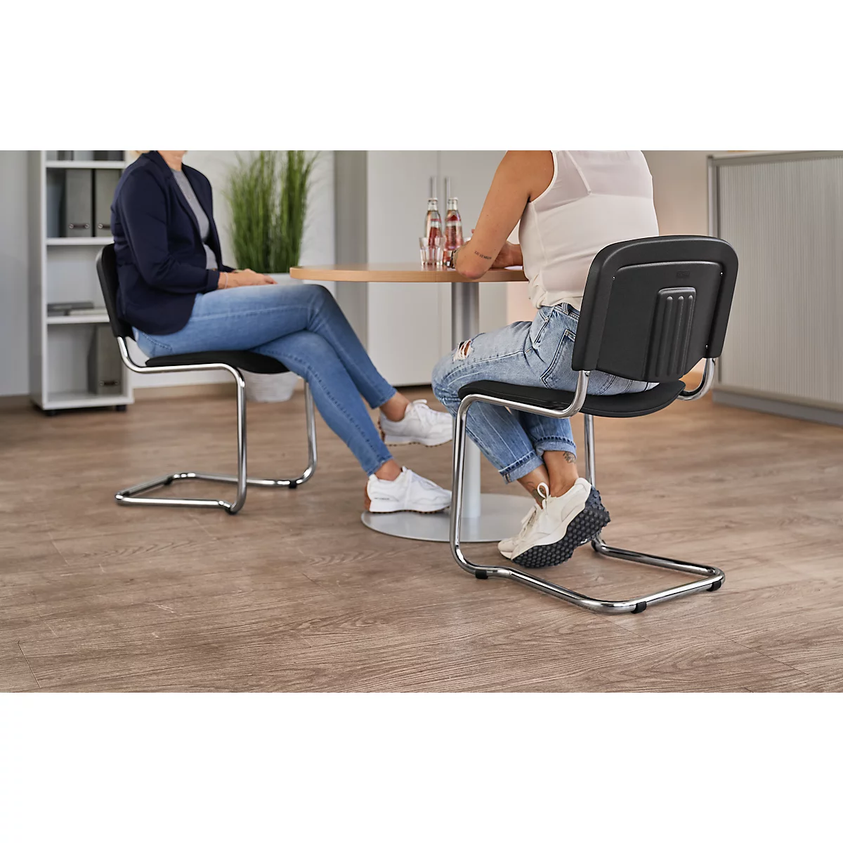 Zwei Personen sitzen an einem Tisch. Der Fokus liegt auf den Stühlen, die ein modernes Design mit schwarzen Rückenlehnen und Metallrahmen aufweisen.