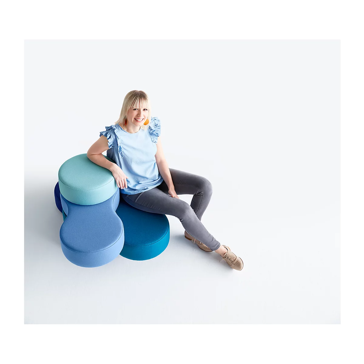 Eine Frau in blauem Oberteil sitzt auf einem blauen, runden Sitzmöbel vor weißem Hintergrund.