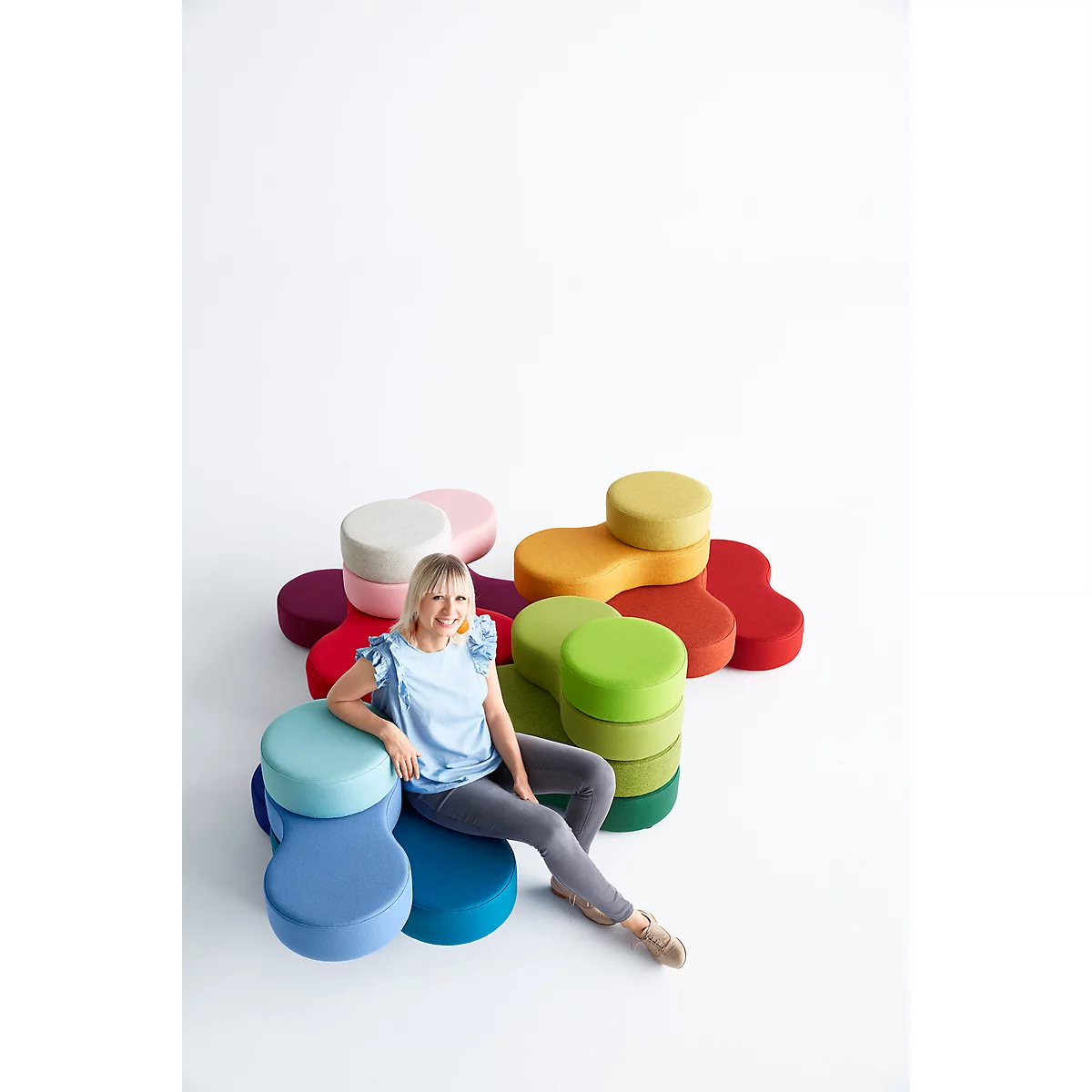 Eine Frau sitzt auf bunten, organisch geformten Hockern. Weitere Hocker liegen im Hintergrund. Sie trägt ein hellblaues Oberteil und graue Hosen.