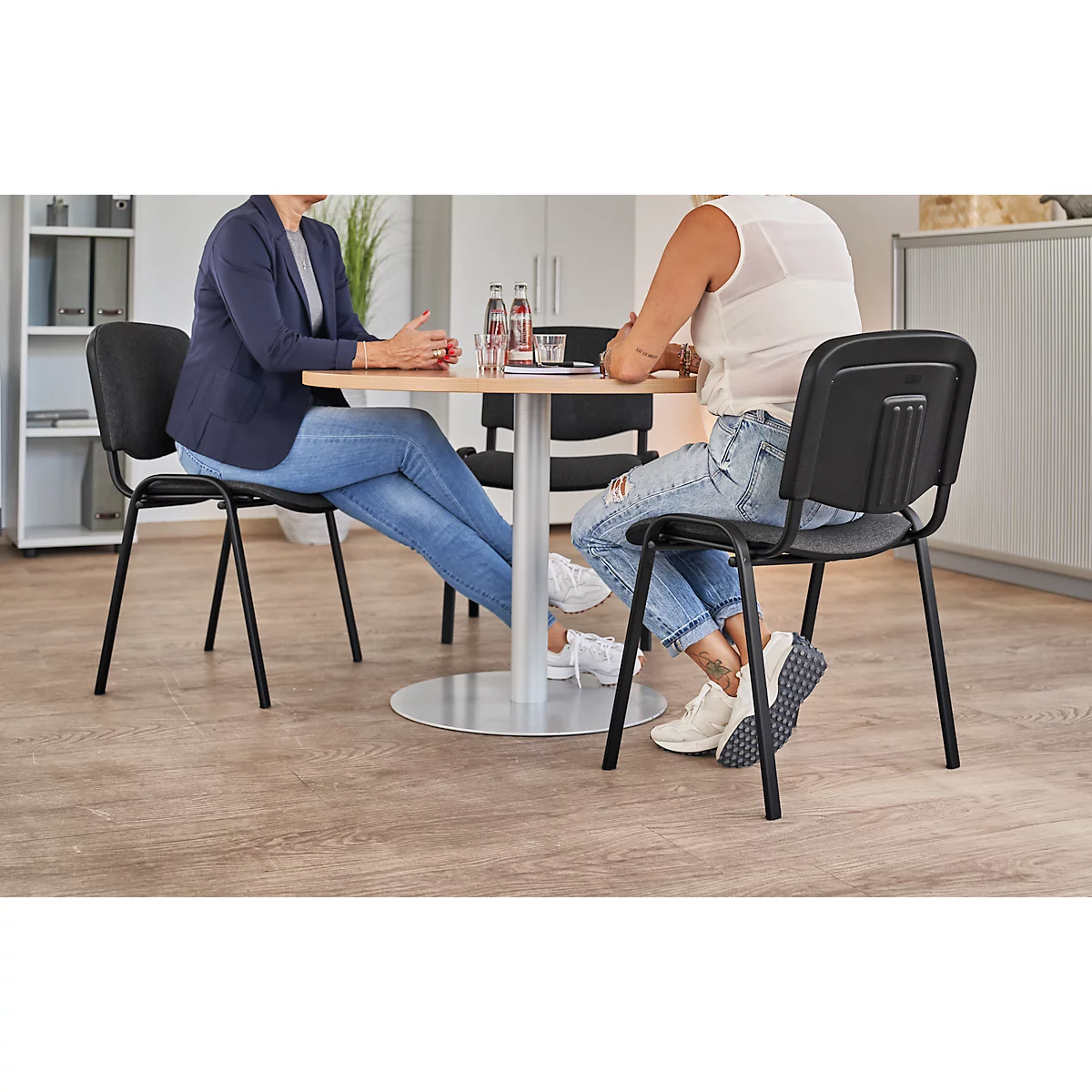 Zwei Frauen sitzen an einem Tisch, sie unterhalten sich. Auf dem Tisch stehen Flaschen und Gläser. Beide tragen Jeans.