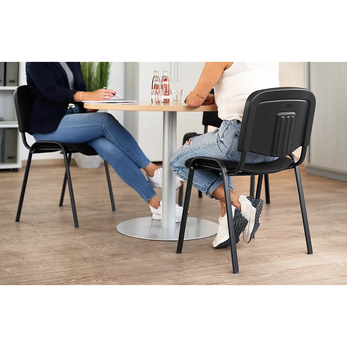 Zwei Personen sitzen an einem runden Tisch, beide tragen Jeans und Turnschuhe. Der Hintergrund ist ein Büro.