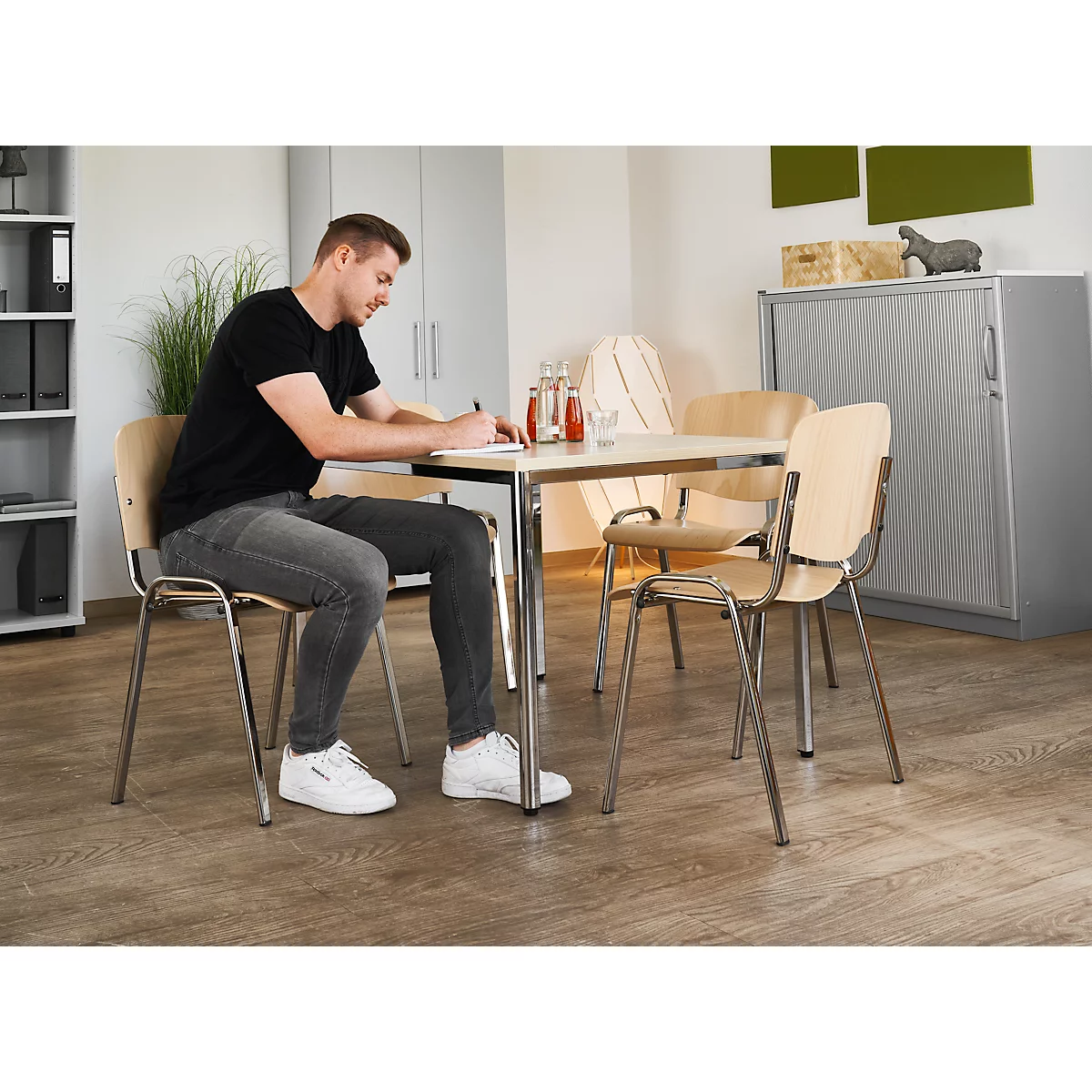 Ein Mann sitzt an einem Tisch und schreibt. Drei weitere Stühle stehen um den Tisch herum. Im Hintergrund ein Schrank.