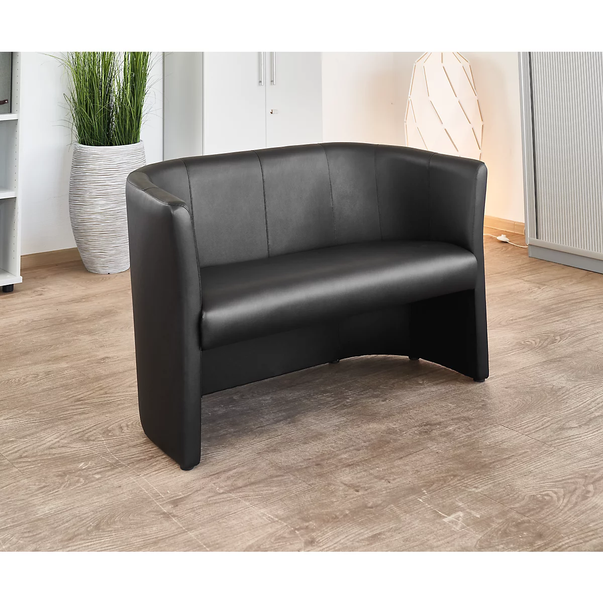 Schwarzes Ledersofa in einem Büro. Es steht auf hellem Holzfussboden. Im Hintergrund ein Topfpflanze.