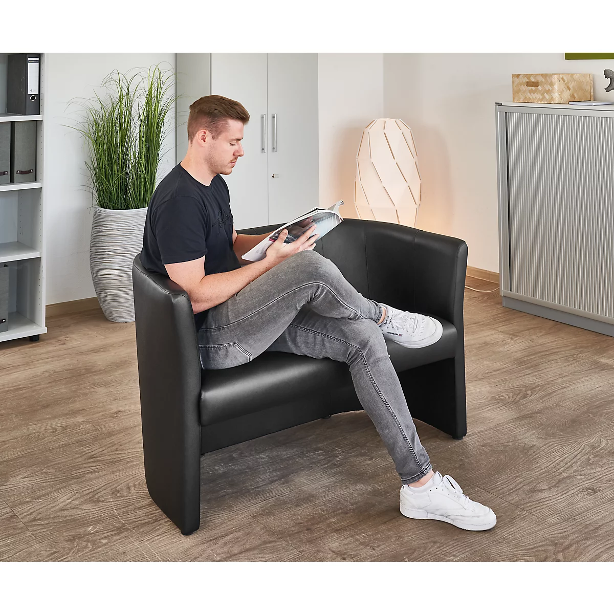 Ein Mann sitzt in einem schwarzen Ledersessel und liest eine Zeitschrift. Er trägt Jeans, ein schwarzes T-Shirt und weisse Schuhe.