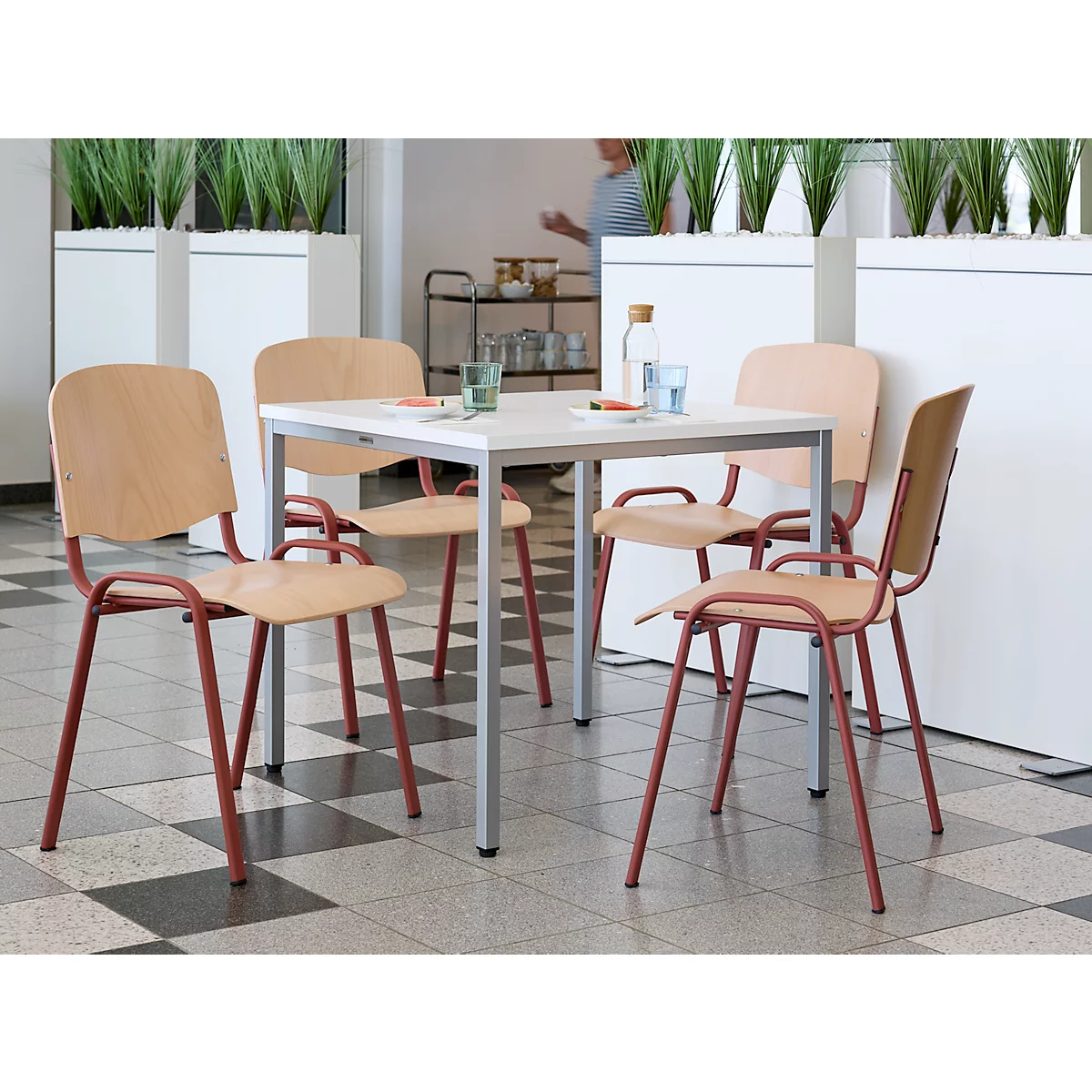 Vier stoelen rond een tafel met glazen, waterfles, persoon op de achtergrond. Houten rugleuningen, roodbruine poten. Lichte omgeving.