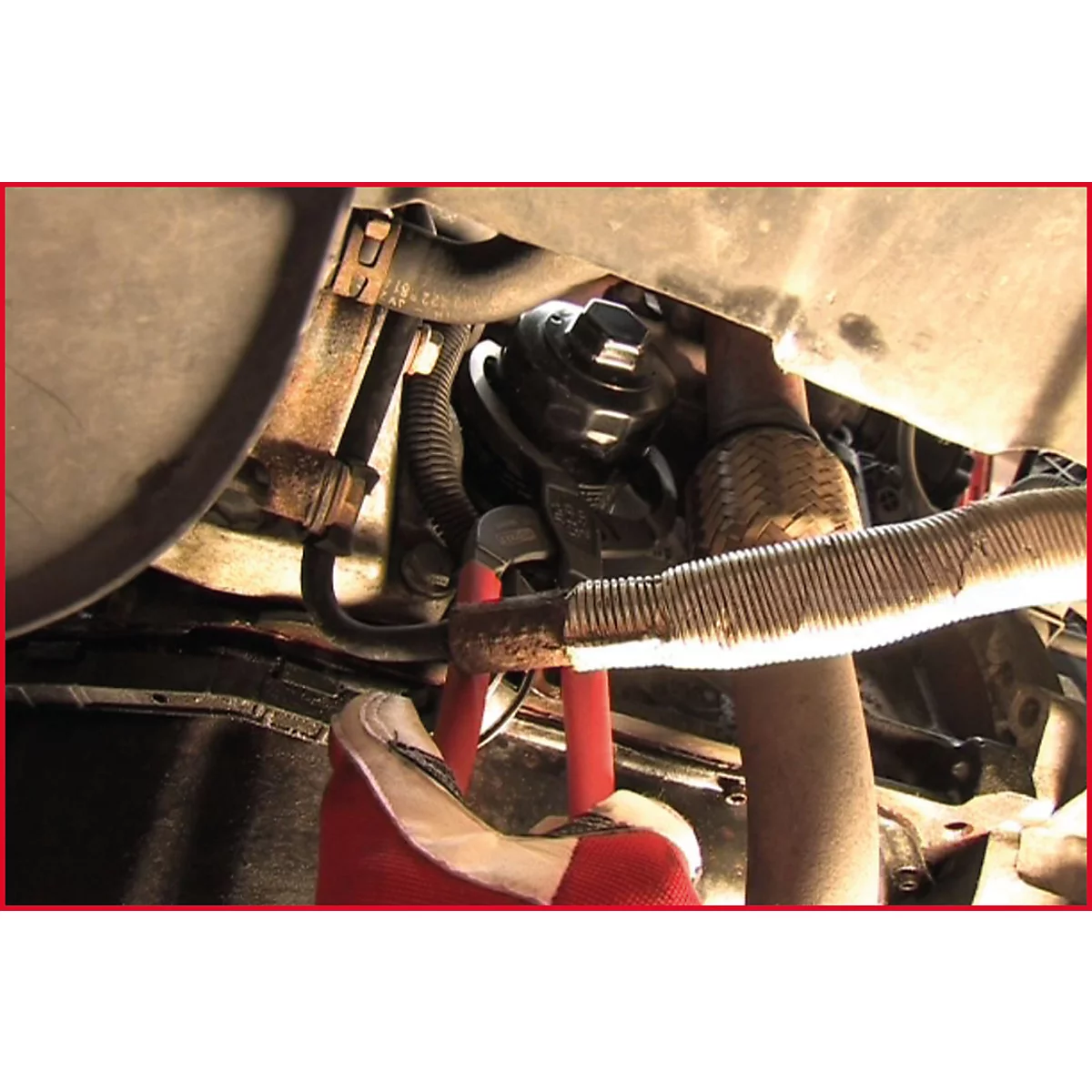 Nahaufnahme eines Motorraums mit Werkzeug und Rohren. Eine Hand in rotem Handschuh hält eine Zange.