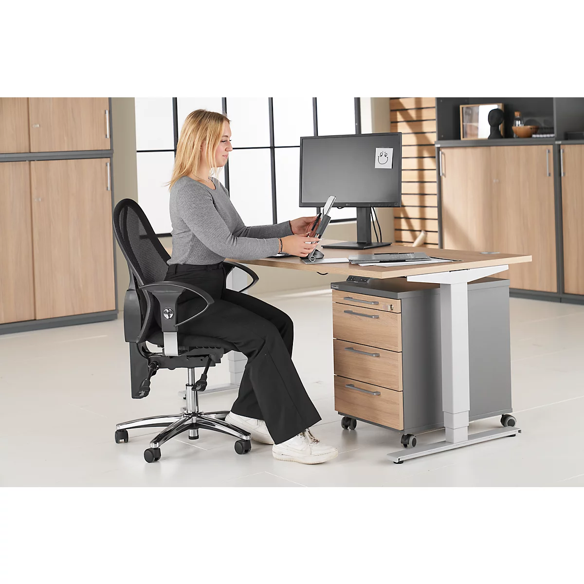 A woman sits at a height-adjustable desk working with a tablet. Next to her is an office chair.