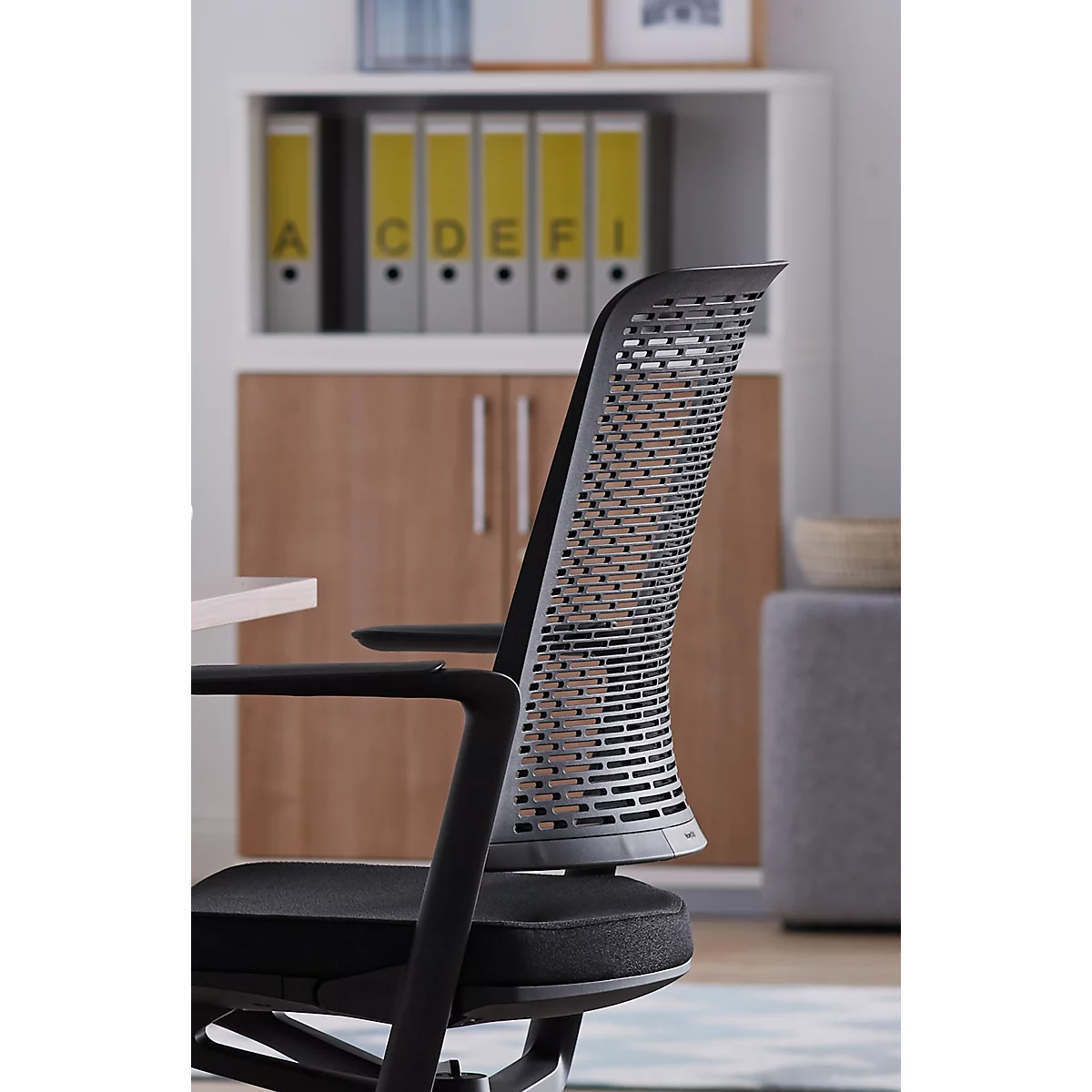 Black office chair in front of white shelf with yellow folders. The chair has a perforated backrest.