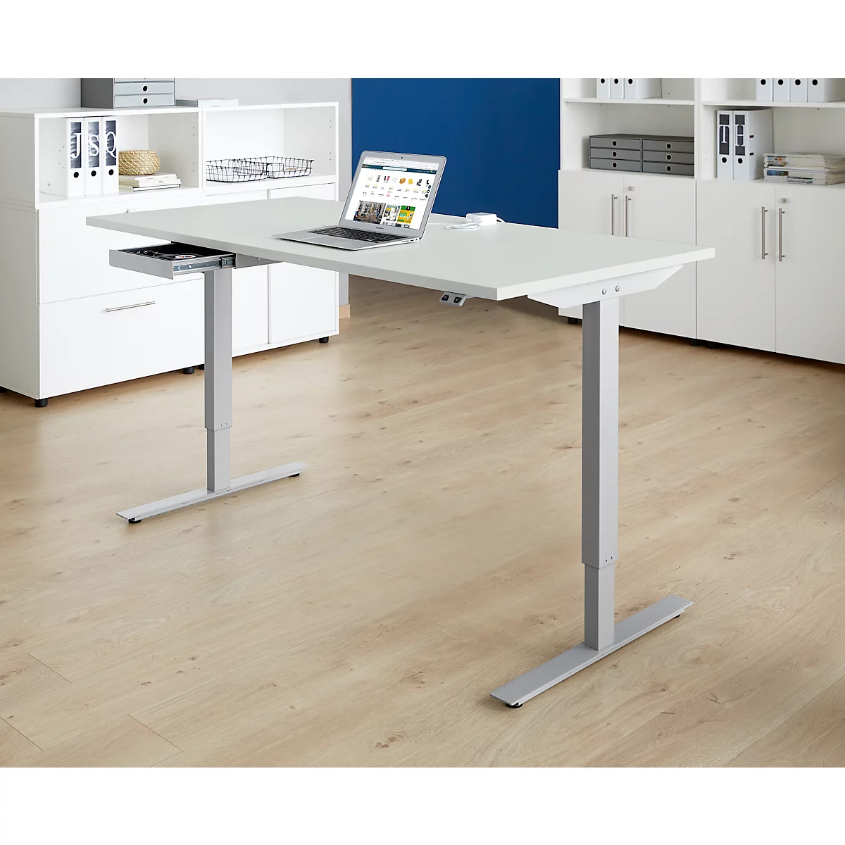 Un bureau blanc réglable en hauteur avec un ordinateur portable et un tiroir ouvert se dresse devant des armoires blanches. Le sol est carrelé clair.