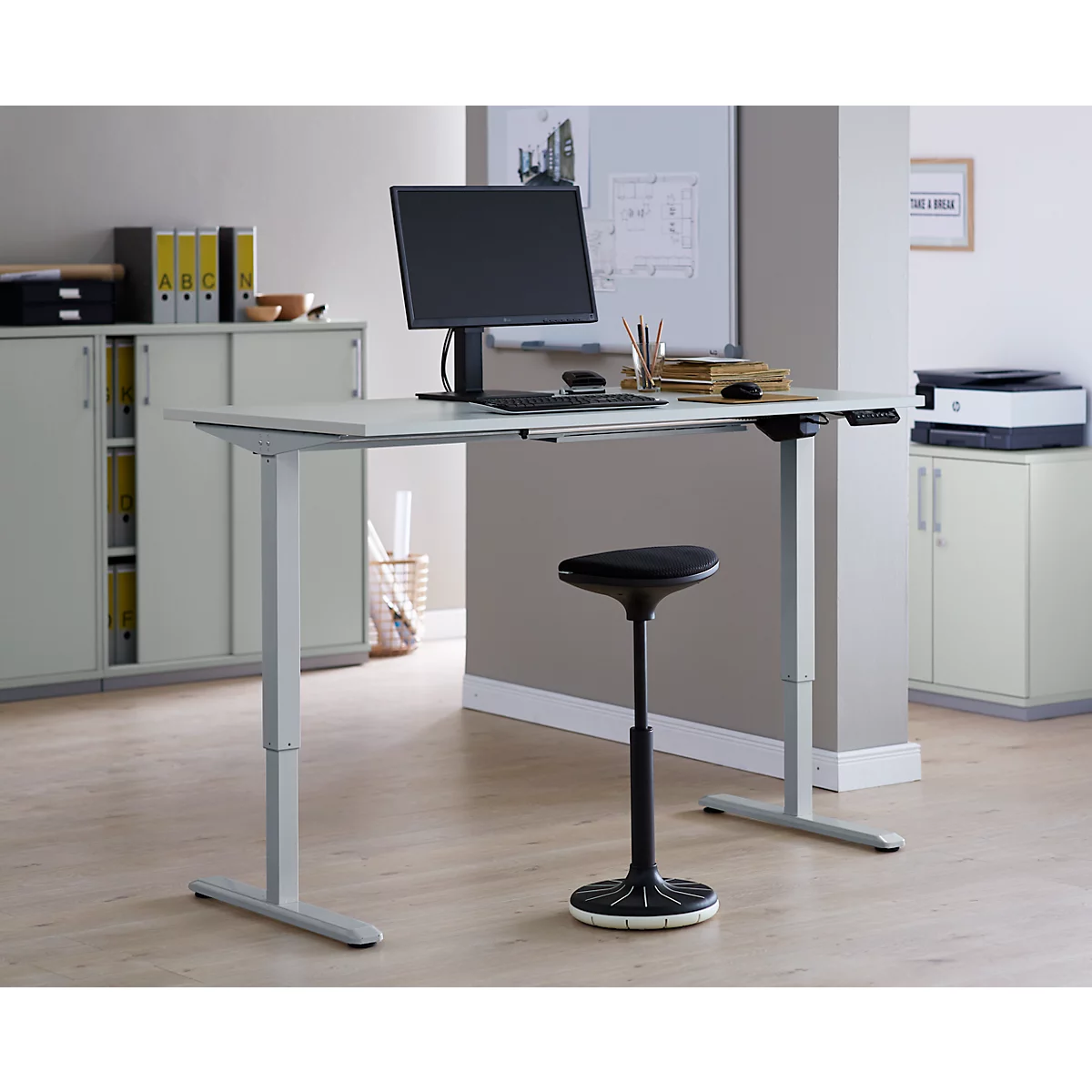 Un bureau réglable en hauteur avec un moniteur et une chaise dans un bureau moderne. Le bureau est gris, la chaise noire.