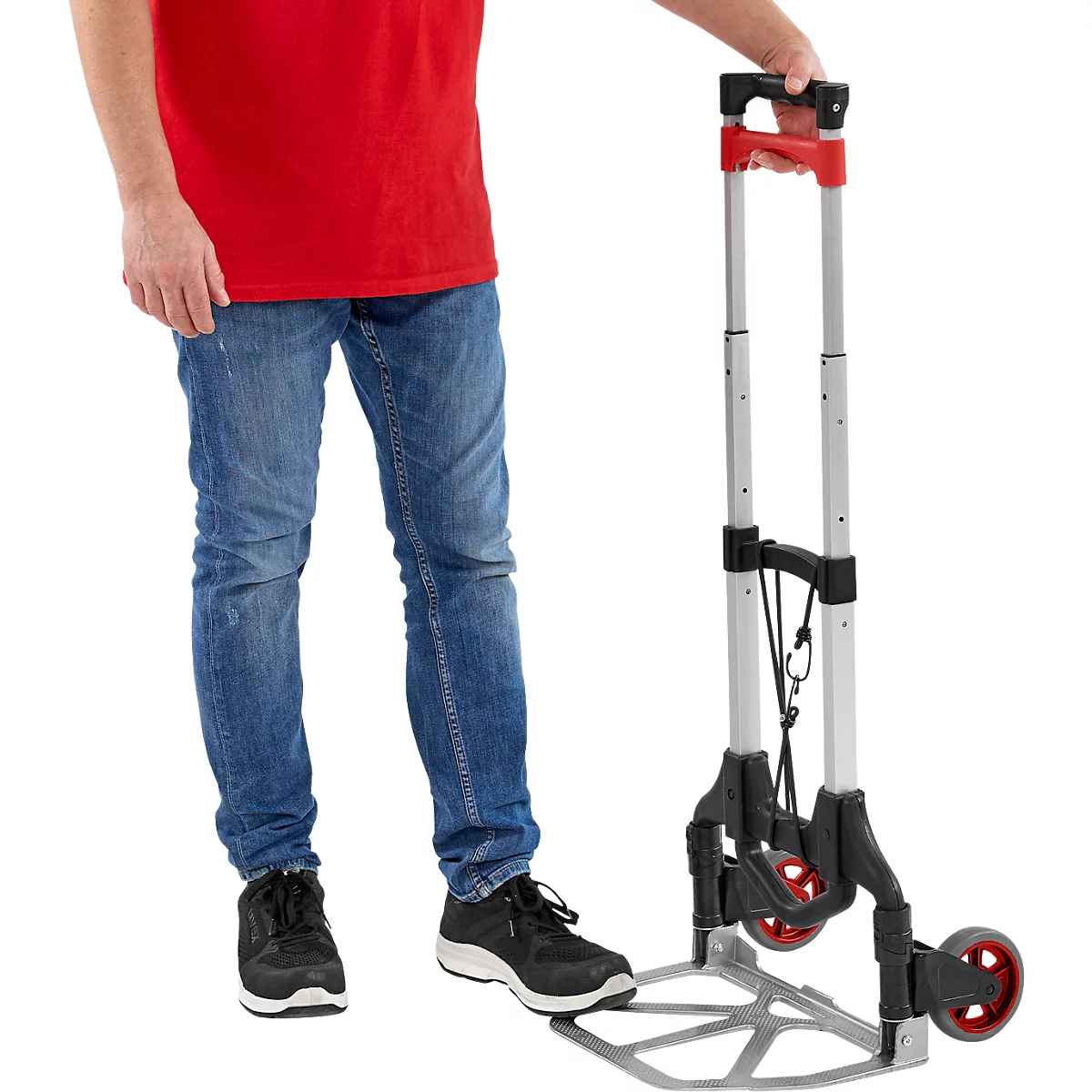 Een persoon houdt een opgevouwen zilveren steekwagen met rode wielen en een rode handgreep, staande op een witte achtergrond.