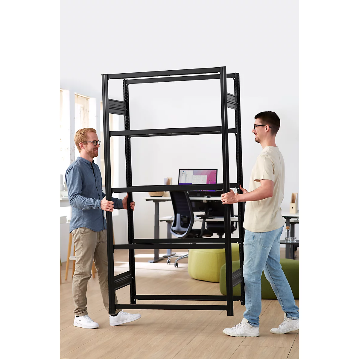 Two men carry a black shelf in an office, they are smiling.