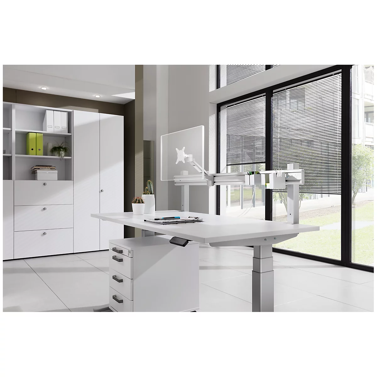 White height-adjustable desk in a bright office. Pens and a monitor are on the table. In the background is a wall unit and window.