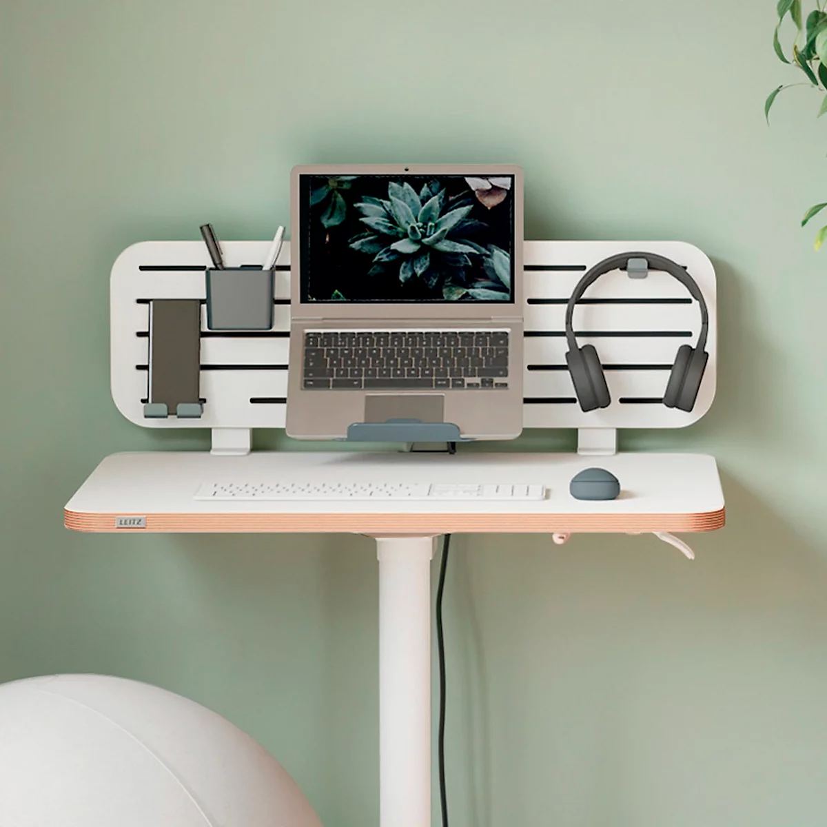 Bureau debout avec ordinateur portable, casque et fournitures de bureau contre un mur vert clair.