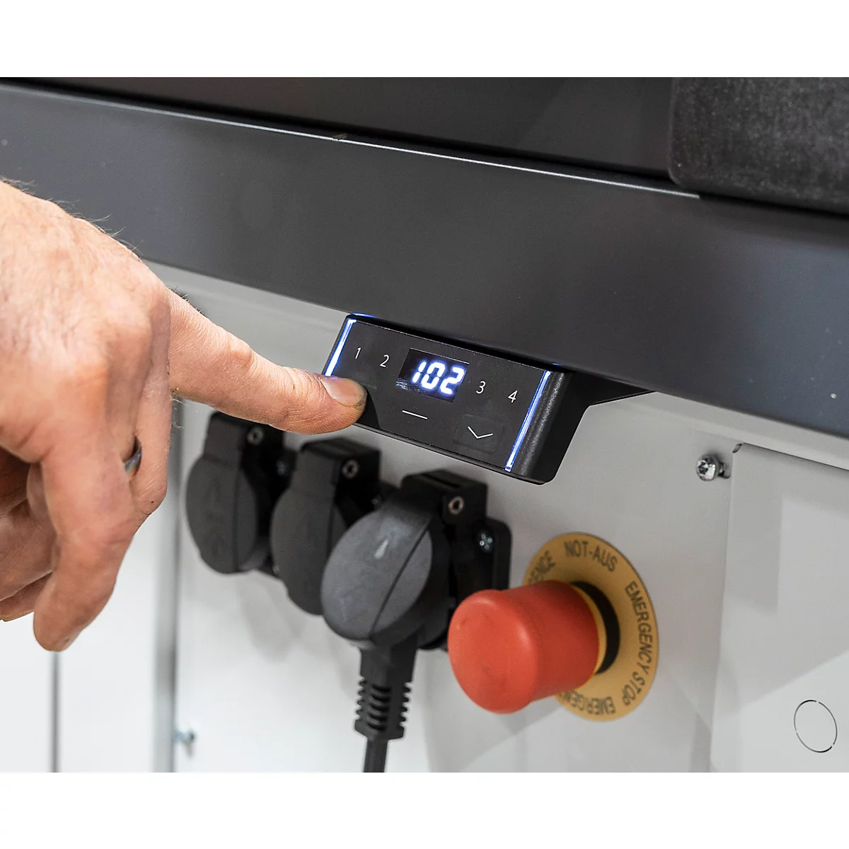 Close-up van een hand die een bedieningspaneel met een digitale display bedient. Hieronder bevinden zich stopcontacten en een rode noodstopknop.