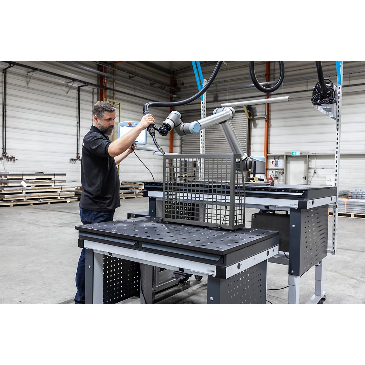 Een man bedient een robotarm die een metalen mand op een tafel inspecteert. Fabriek op de achtergrond.