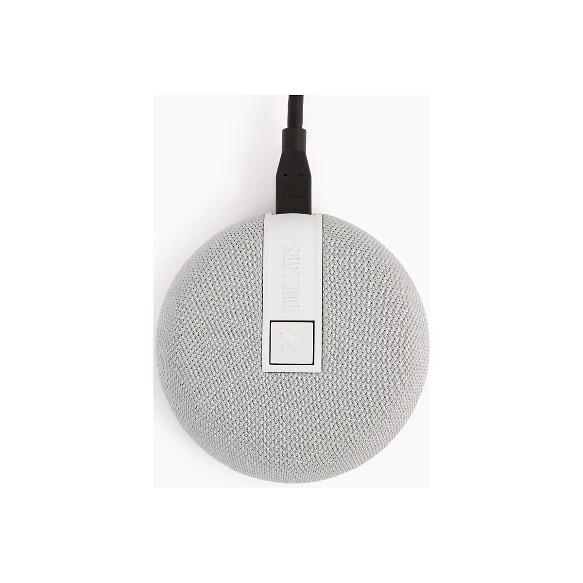 Haut-parleur gris et rond avec surface textile, relié à un câble noir. Sur une surface blanche.