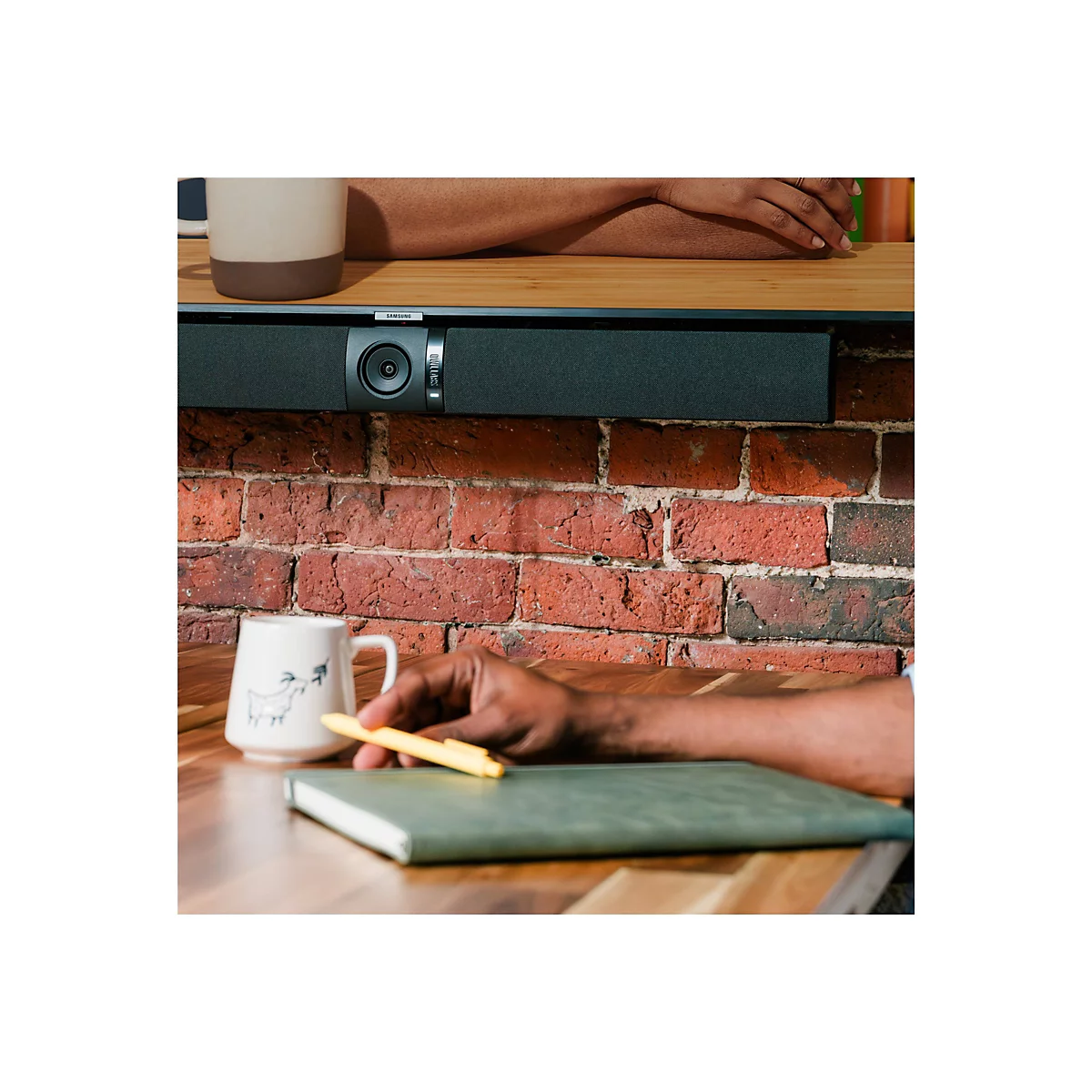Nahaufnahme: Eine Hand hält einen gelben Stift über einem grünen Notizbuch. Eine Tasse und eine Soundbar sind zu sehen.