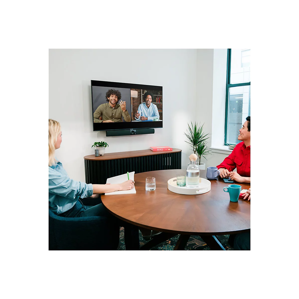 In einem Raum sitzen Personen um einen runden Tisch. Auf einem Fernseher sind zwei Personen in einer Videokonferenz zu sehen.