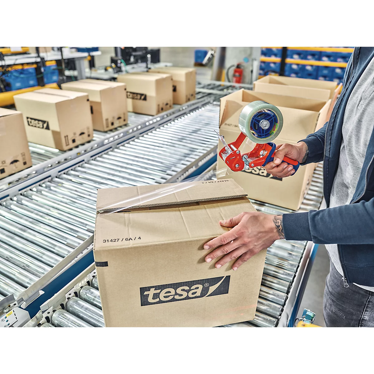 Person verklebt Karton auf Förderband mit tesafilm. Weitere Kartons auf dem Band.