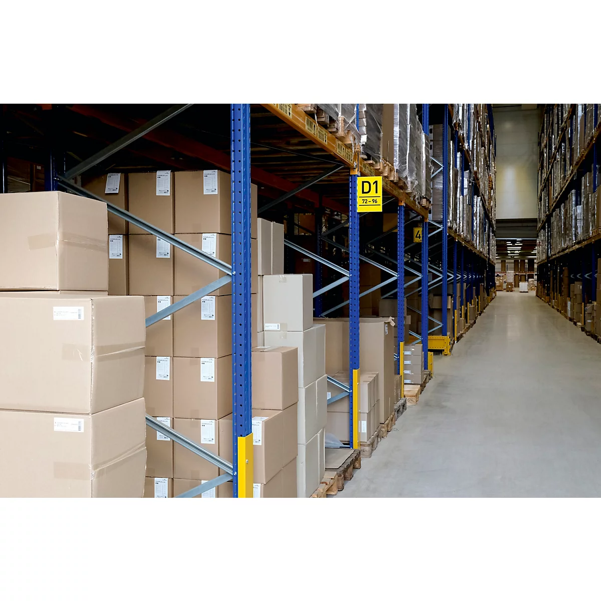 Rangées de boîtes en carton dans un rayonnage d'entrepôt. Un couloir en arrière-plan. D'autres étagères peuvent être vues à droite.
