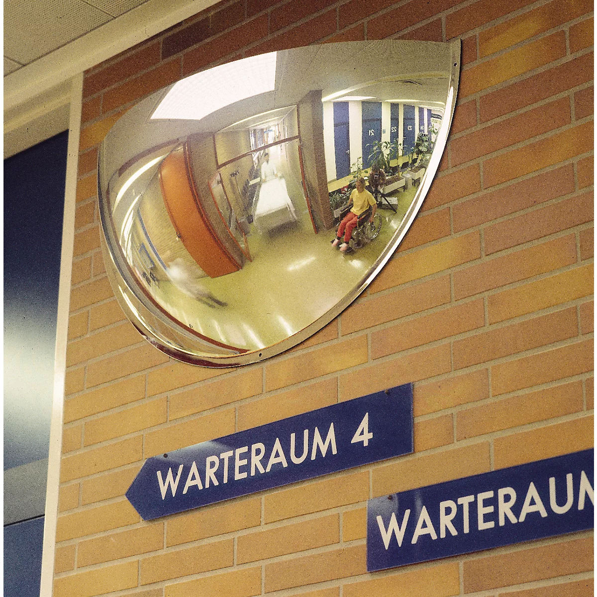 Ein halbkreisförmiger Spiegel an einer Backsteinwand über blauen Schildern mit der Aufschrift Warteraum.