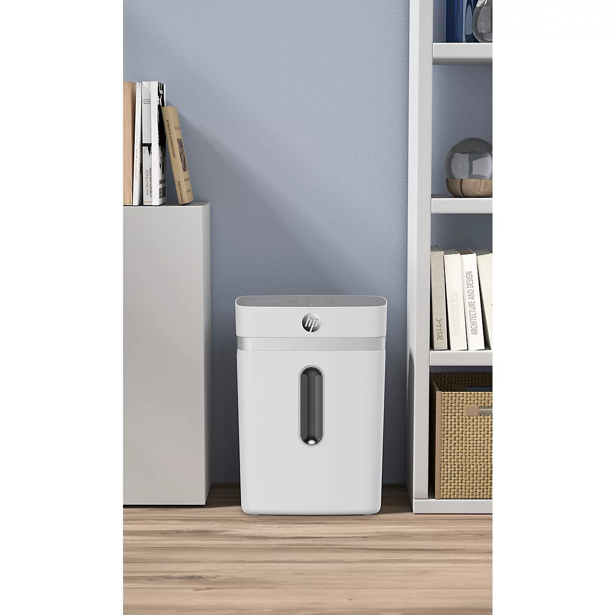 White paper shredder with viewing window, standing on light wooden floor in front of light blue wall. Books to the left, shelf with books to the right.