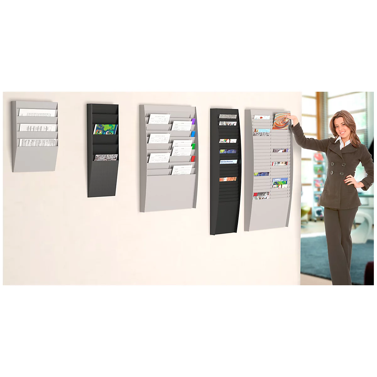 A woman stands beside several wall-mounted brochure holders. Various sizes and colors, filled with brochures and papers.
