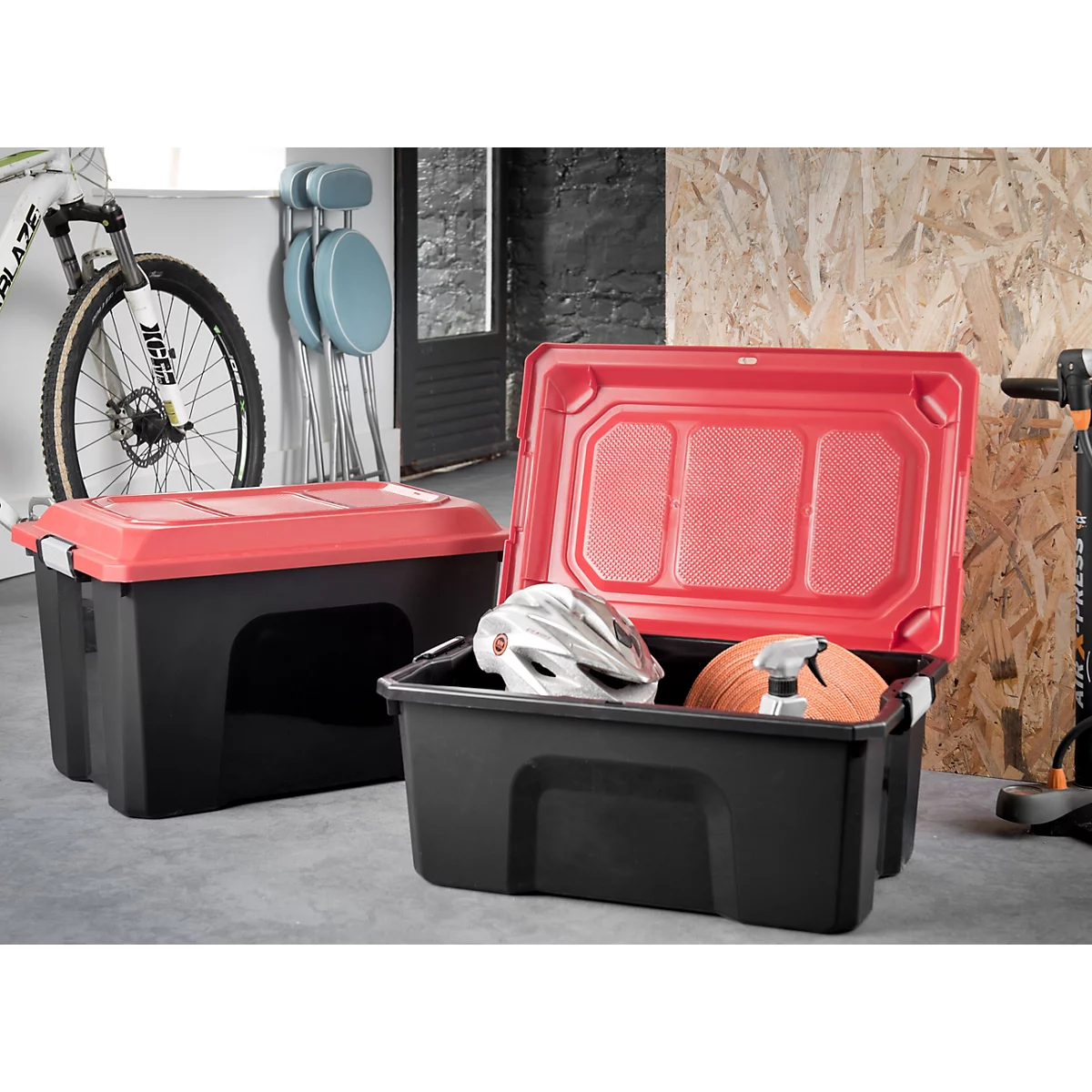 Zwei schwarze Kunststoffboxen mit roten Deckeln. Eine ist geöffnet und enthält Helme. Im Hintergrund ein Fahrrad und Hocker.