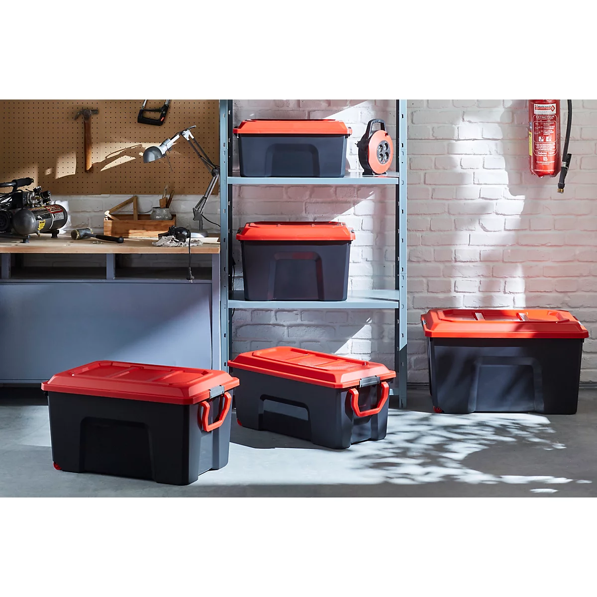 Schwarze Werkzeugboxen mit roten Deckeln stehen in einer Werkstatt. Einige stehen auf einem Regal, andere auf dem Boden.