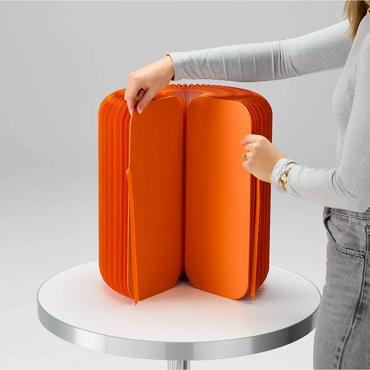 Eine Person faltet einen orangefarbenen, ziehharmonikaartigen Hocker auf einem weißen Tisch auf. Das Design ist klar und modern.