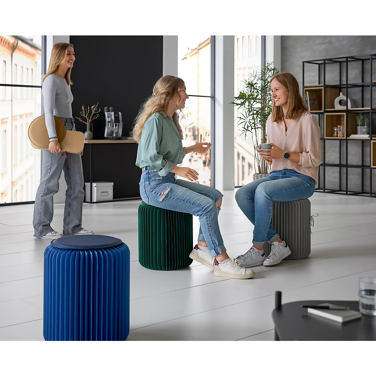 Drie jonge vrouwen zitten en staan in een moderne kamer. Ze praten met elkaar, een van de vrouwen houdt een kruk vast.