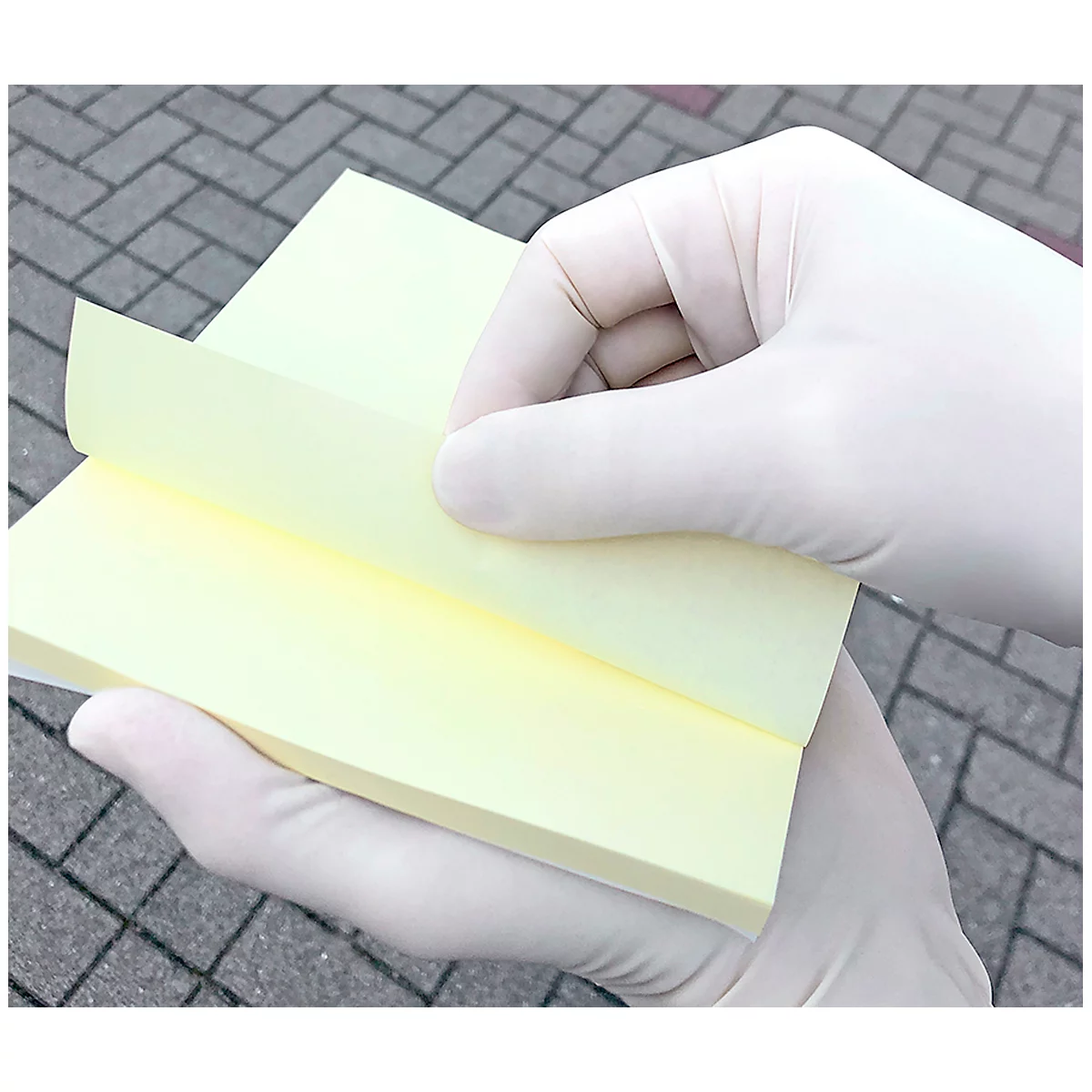Des mains gantées de blanc soulèvent un feuillet jaune. En arrière-plan, un chemin pavé est visible.