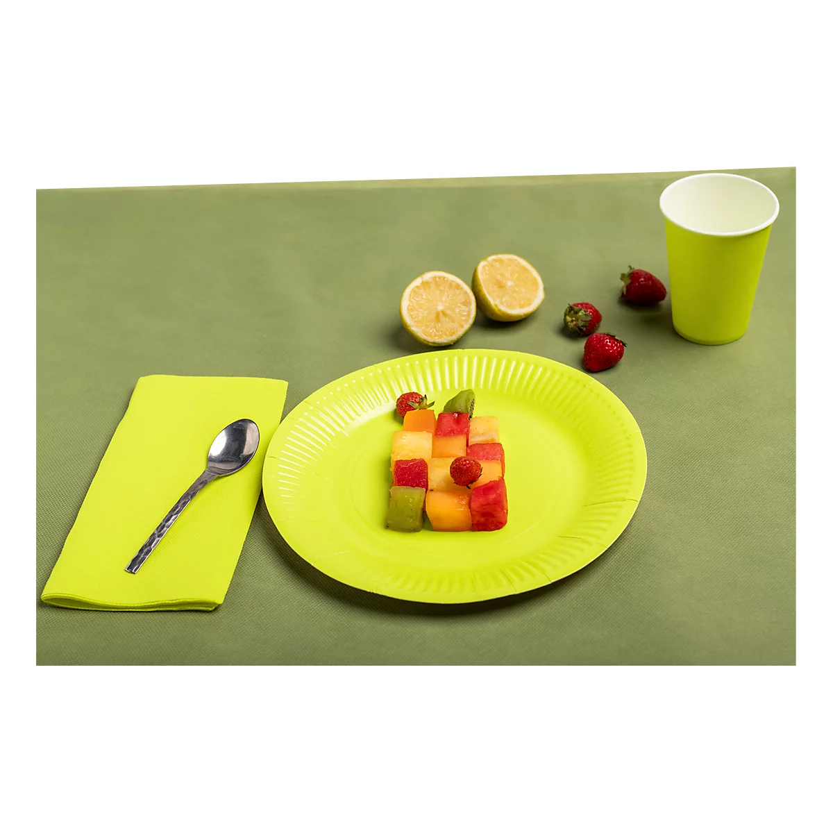 Teller mit bunt gewürfeltem Obst, Zitrone, Erdbeeren, Servietten, Löffel und Becher auf grünem Untergrund.