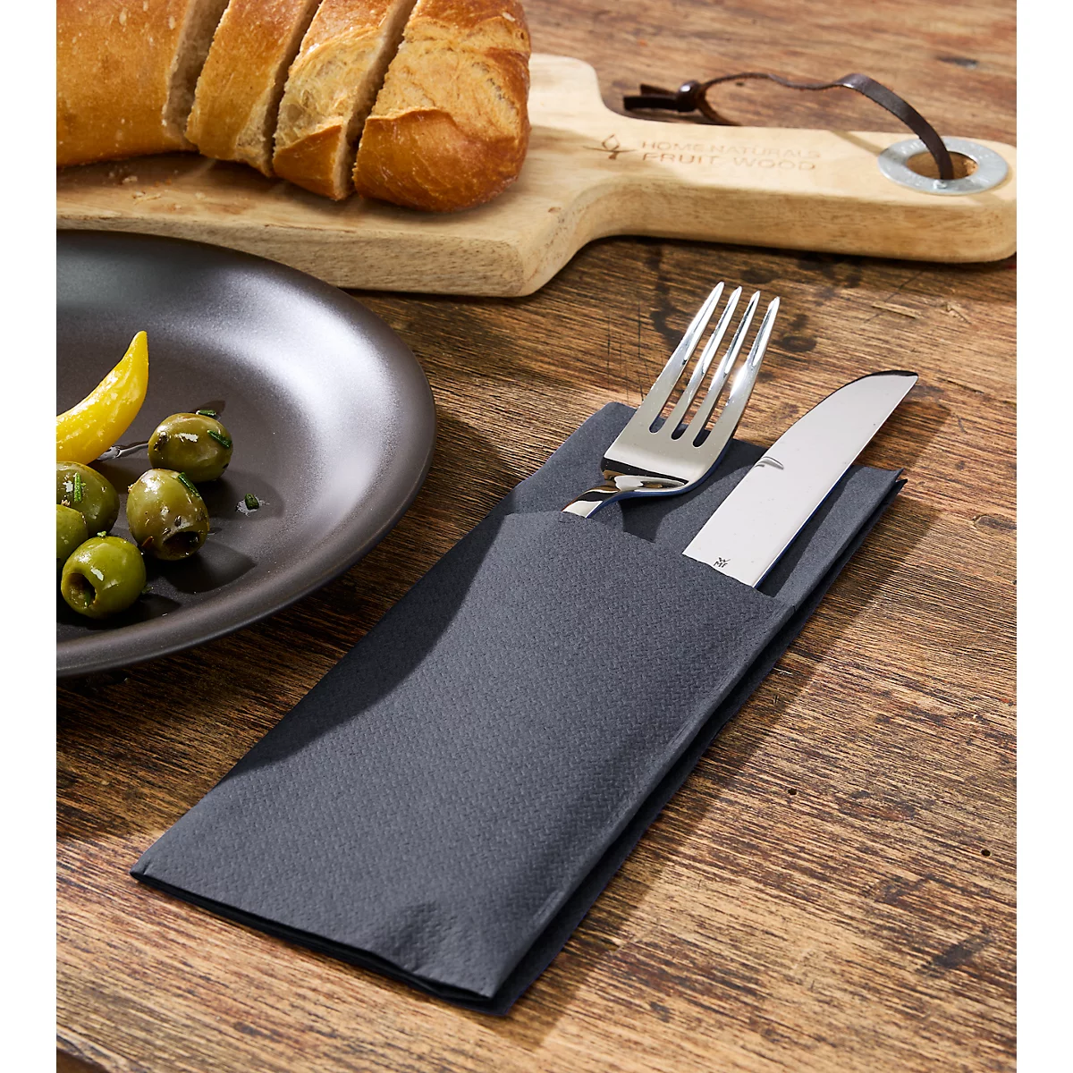 Besteck in grauer Serviette auf Holztisch. Links Teller mit Oliven und Zitrone, Brot und Schneidebrett im Hintergrund.