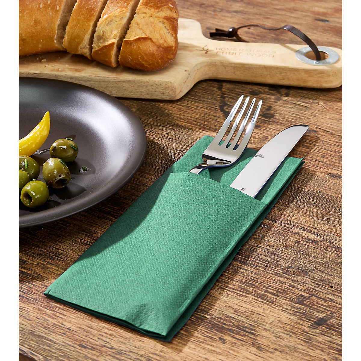 Bestek in groene servet op houten tafel. Bord met olijven links, brood en snijplank op de achtergrond.